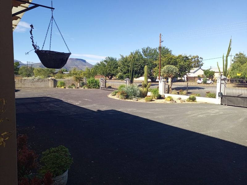 A spacious paved driveway leading to a secure gated entrance, featuring landscaped garden beds and mountain views in the background.