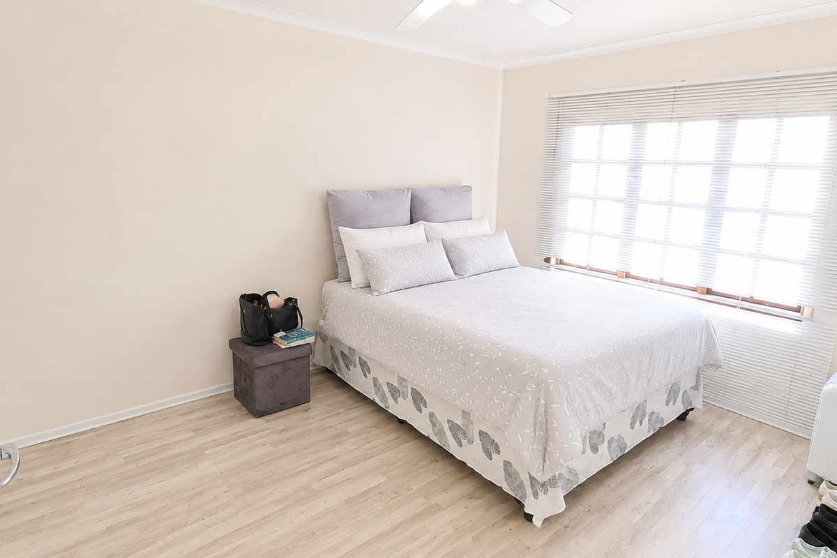 A bright and neutral bedroom featuring light laminate flooring, a ceiling fan, and large windows fitted with venetian blinds.