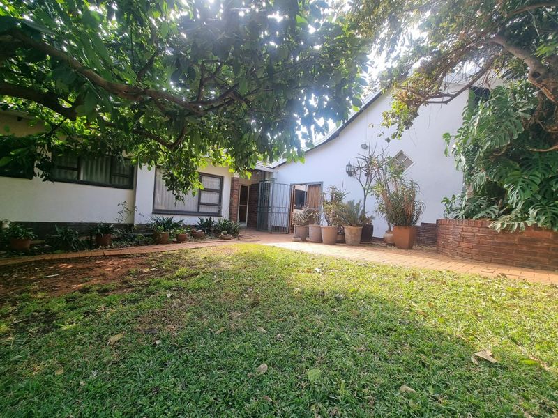 A rear garden area featuring brick paving, a built-in brick planter, and mature trees providing shade against the house exterior.