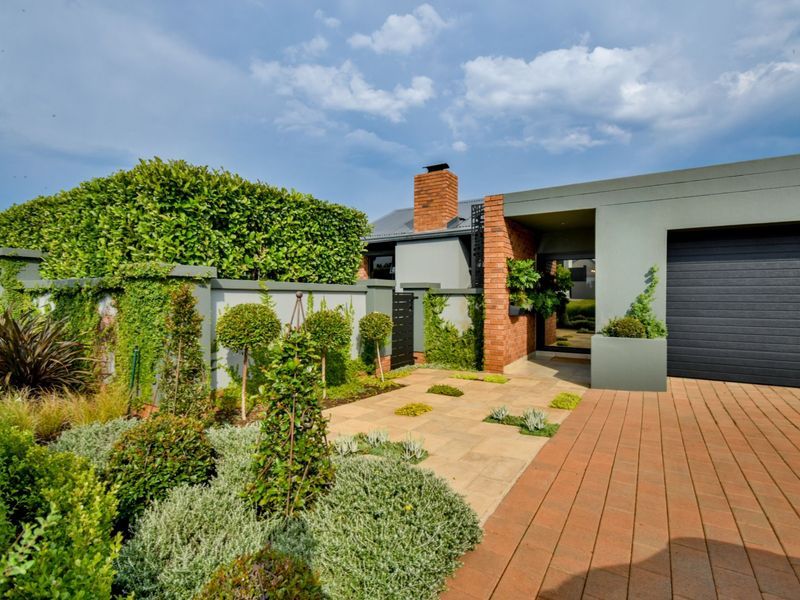A well-maintained front exterior featuring a modern facade, brick chimney, paved driveway, automated garage door, and professionally landscaped garden.