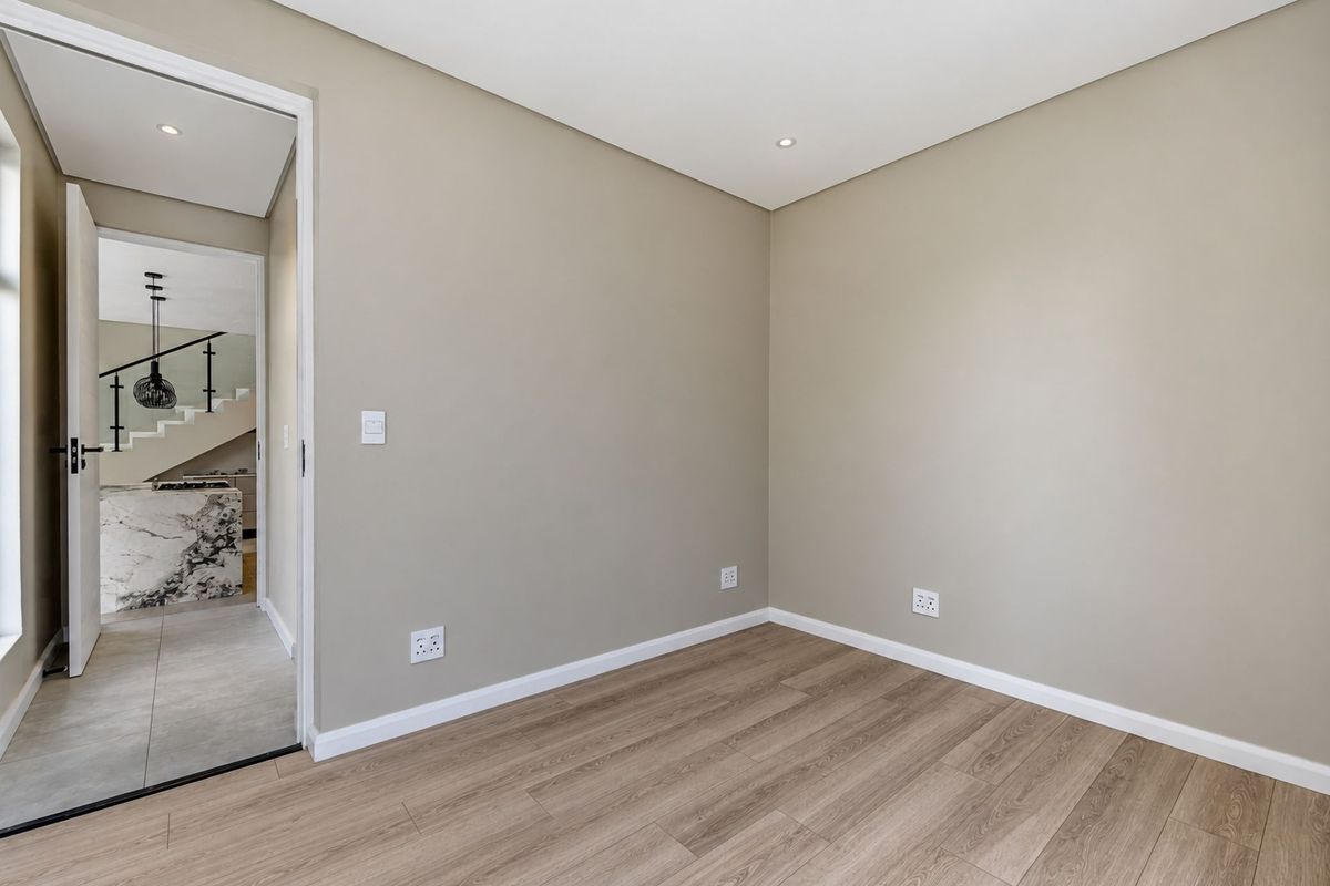 A bright, empty bedroom featuring modern laminate flooring, neutral wall tones, and recessed lighting, with a view into the adjacent hallway and staircase area.