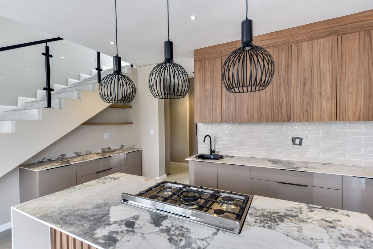 Modern kitchen featuring stone countertops, gas cooktop, wood-finish cabinetry, and subway tile backsplash, adjacent to a contemporary staircase with glass balustrade.