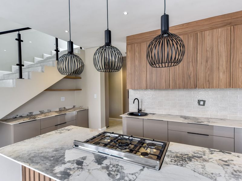 Modern kitchen featuring stone countertops, gas cooktop, wood-finish cabinetry, and subway tile backsplash, adjacent to a contemporary staircase with glass balustrade.