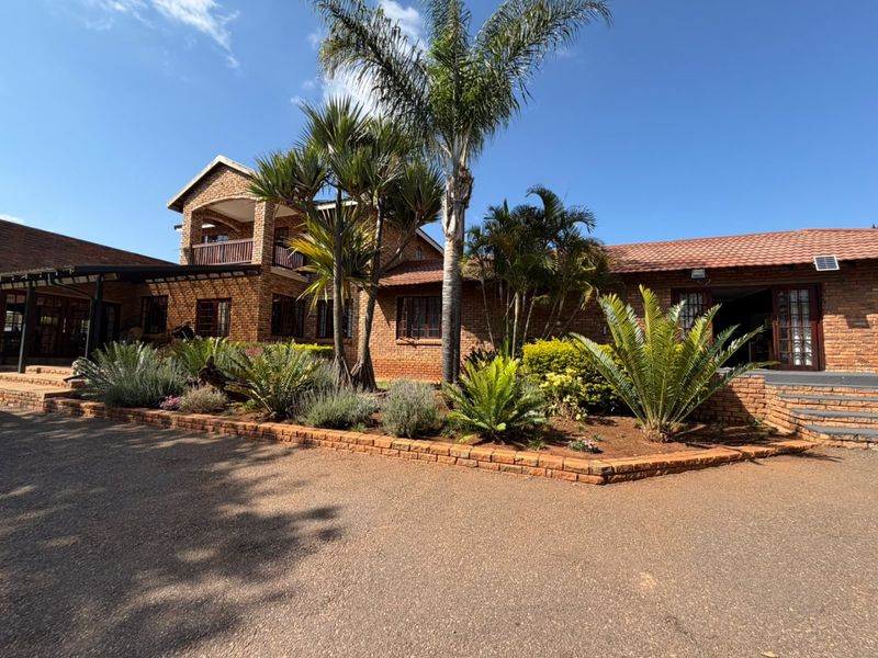A well-maintained face brick farm-style residence featuring a tiled roof, covered balcony, and established garden beds along a paved driveway.