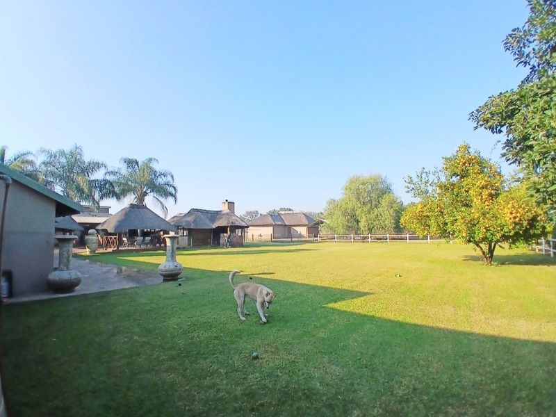 Spacious farm garden featuring a large lawn, mature trees, and multiple thatched-roof outdoor structures with a built-in fireplace.