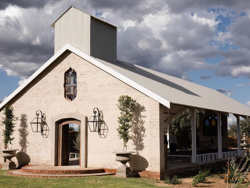 A charming chapel-style building featuring a brick facade, corrugated metal roof, arched window, and a covered patio area, set within a landscaped garden.