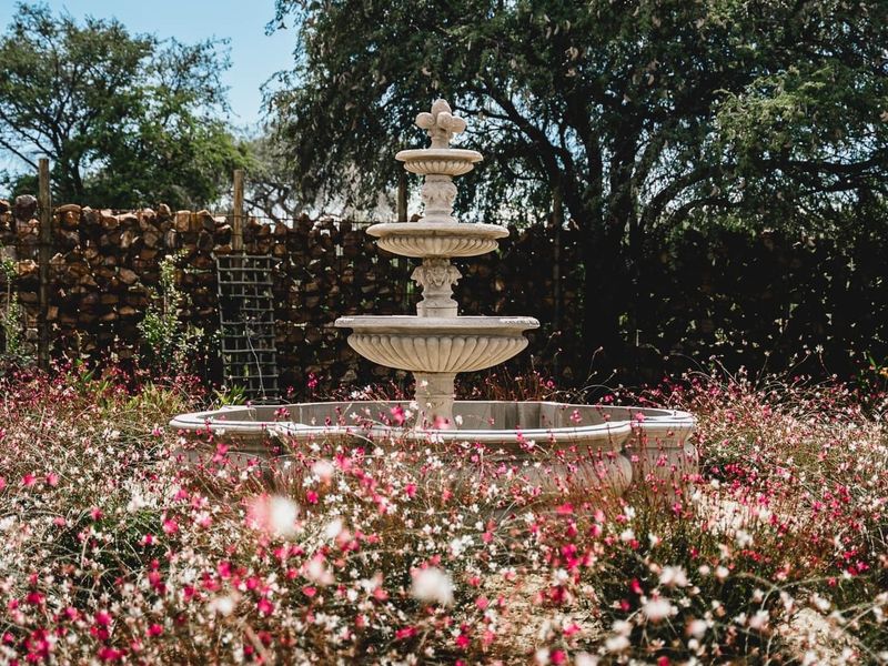 A decorative tiered stone fountain set within a lush, flowering garden, framed by a rustic stone boundary wall.