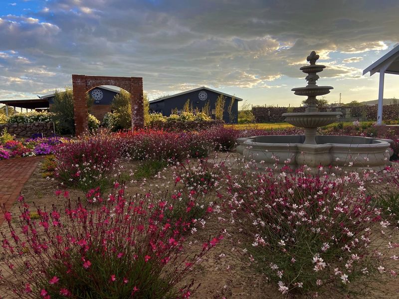 A beautifully landscaped garden featuring a central stone fountain, a decorative brick archway, and established flower beds with a brick pathway.