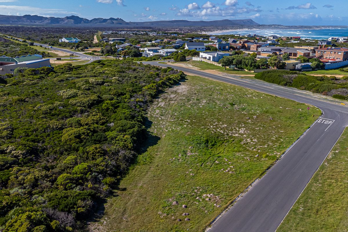 An aerial view of a vacant residential plot situated in a coastal neighborhood, featuring surrounding vegetation and proximity to local roads and the coastline.