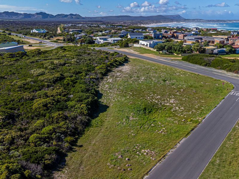 An aerial view of a vacant residential plot situated in a coastal neighborhood, featuring surrounding vegetation and proximity to local roads and the coastline.