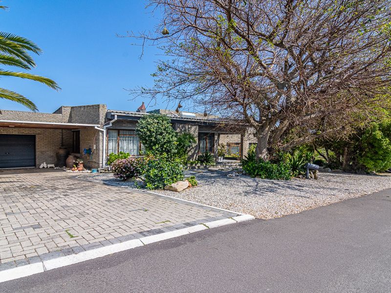 Front exterior view of a single-story brick house featuring a paved driveway, attached garage, carport, and established garden with a large mature tree.