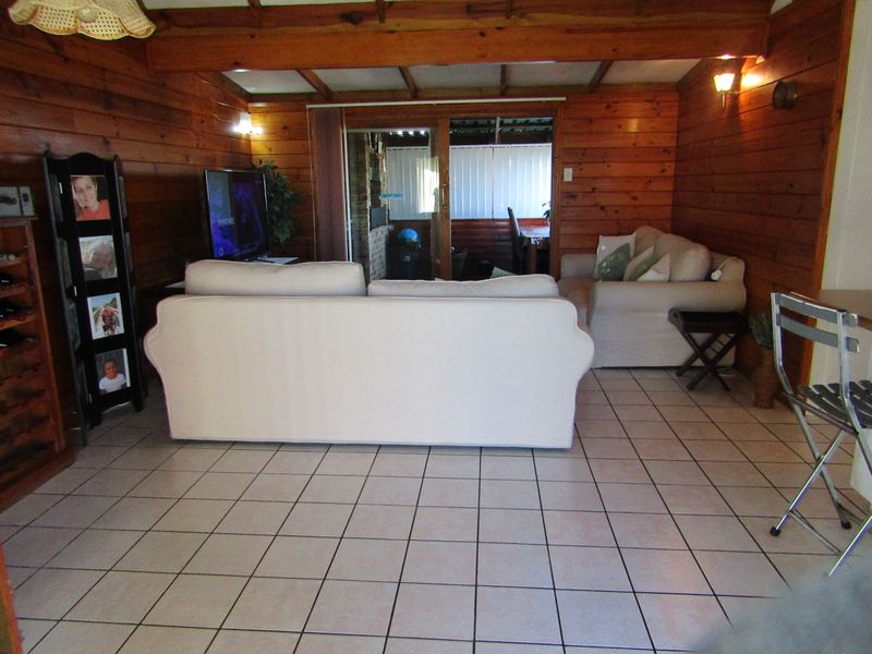 A living room featuring warm timber wall paneling, exposed ceiling beams, and ceramic tiled flooring, with a sliding glass door leading to an outdoor area.