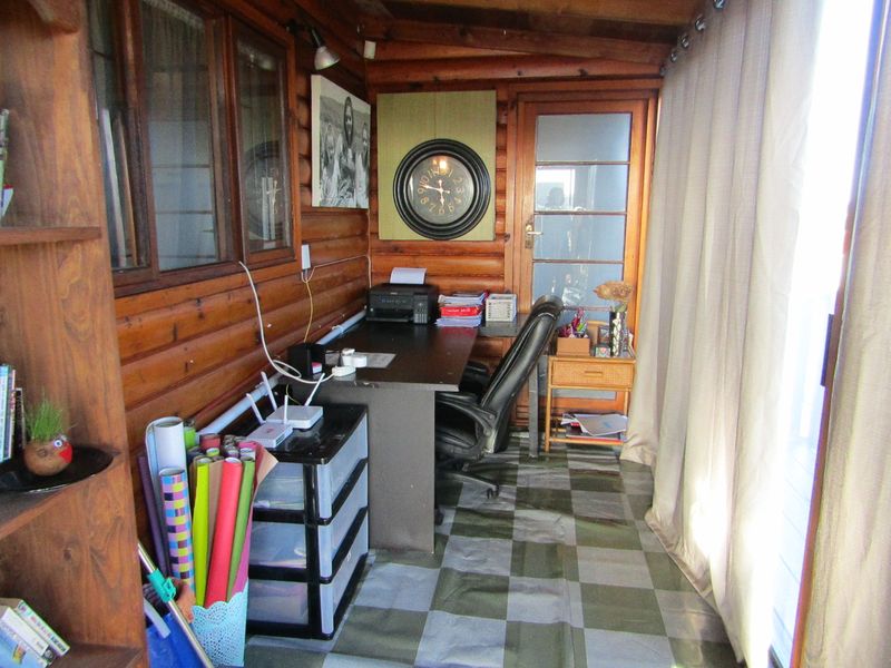 A compact home office space featuring timber log walls, tiled flooring, and natural light through timber-framed windows and a glass door.