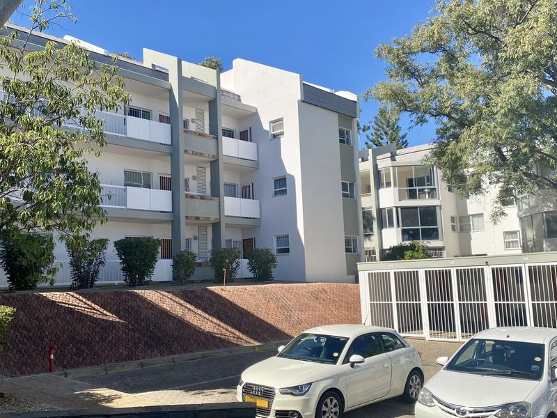 Exterior view of a multi-story apartment complex featuring balconies, a paved parking area, and secure gated access.
