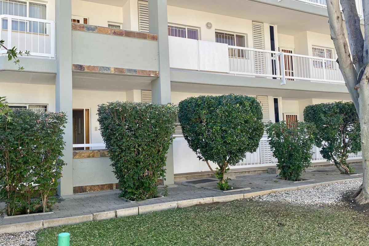 Rear elevation of an apartment building featuring private balconies with railings, stone accents, and a paved ground-level walkway adjacent to a landscaped garden area.