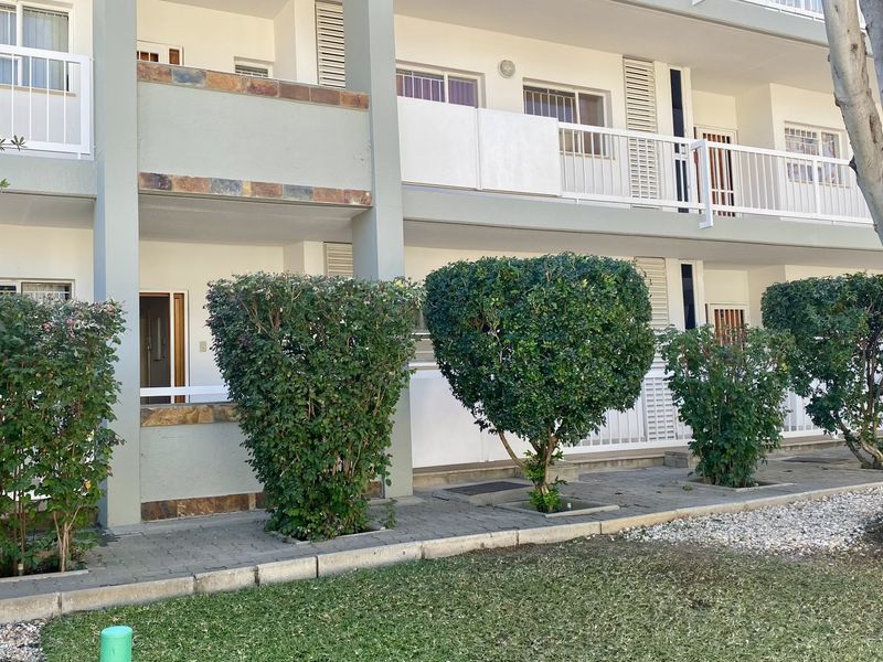 Rear elevation of an apartment building featuring private balconies with railings, stone accents, and a paved ground-level walkway adjacent to a landscaped garden area.