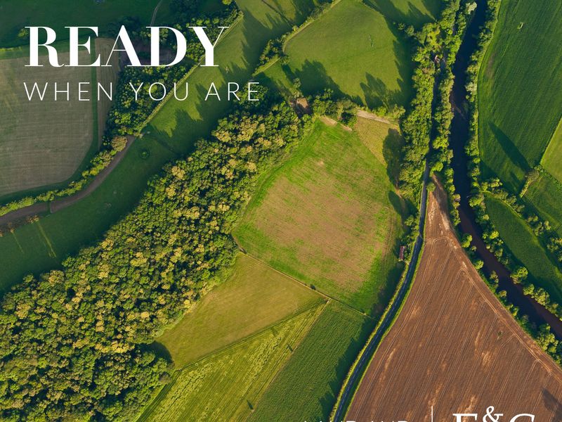 An aerial view of expansive rural land featuring open fields, mature woodland, and a bordering river.