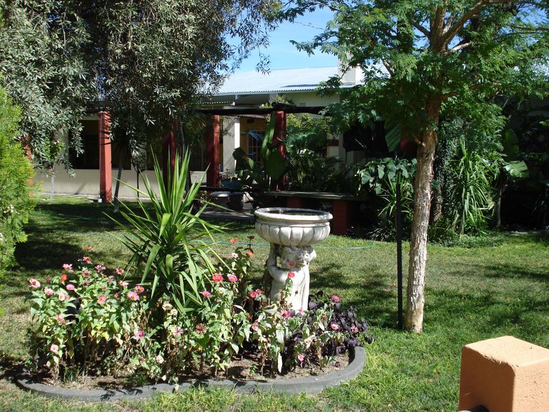 A lush garden area featuring a stone birdbath, established trees, and a covered patio with structural pillars, providing a pleasant outdoor living space.