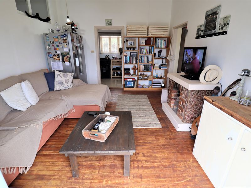 A living room featuring hardwood flooring and a rustic exposed brick fireplace, with an open doorway leading to the kitchen area.