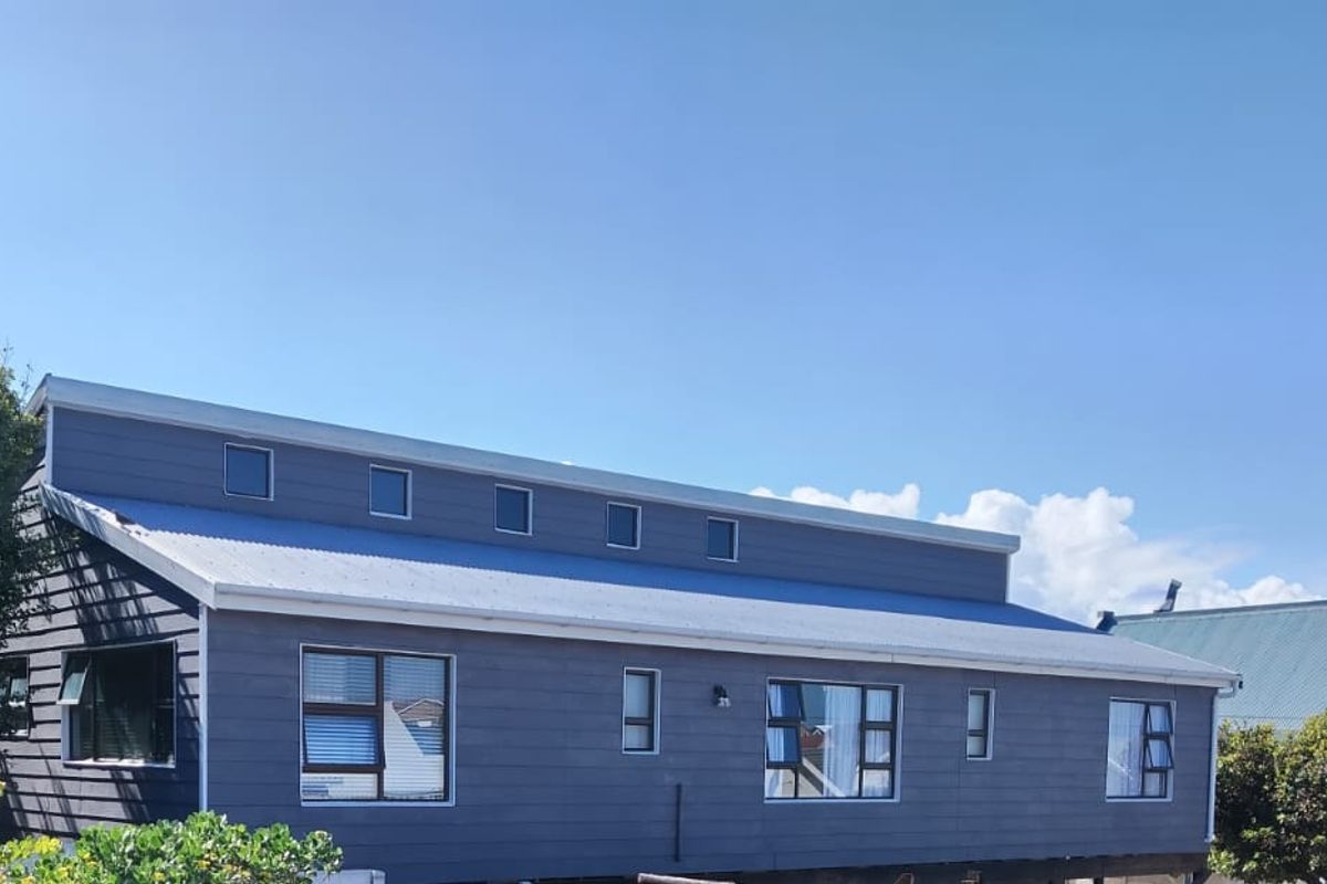 A side view of a modern house featuring dark timber cladding, a metal roof, and multiple aluminium-framed windows including clerestory windows.