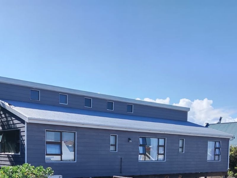 A side view of a modern house featuring dark timber cladding, a metal roof, and multiple aluminium-framed windows including clerestory windows.