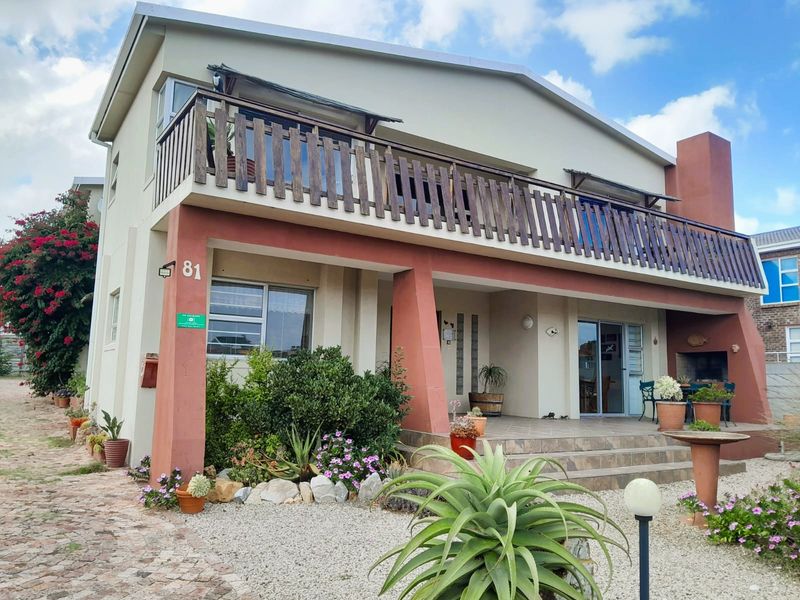 Front elevation of a two-story house featuring a spacious tiled patio, built-in braai, and an upper-level balcony with wooden balustrades.