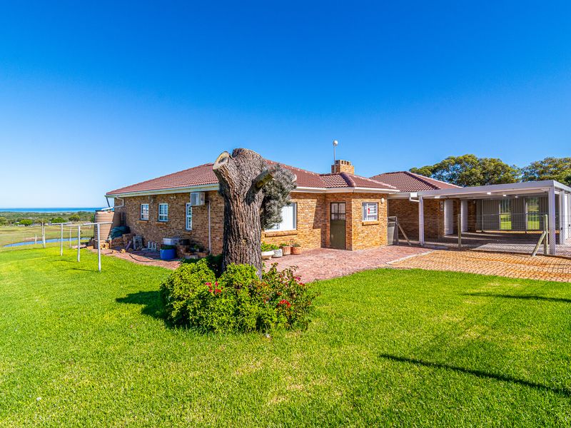 Rear view of a brick farmhouse featuring a paved patio area, attached carport, and expansive lawn with distant landscape views.