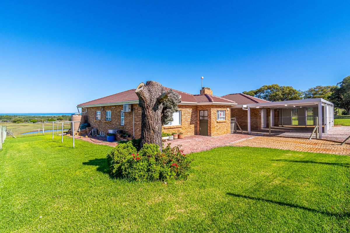 Rear view of a brick farmhouse featuring a paved patio area, attached carport, and expansive lawn with distant landscape views.