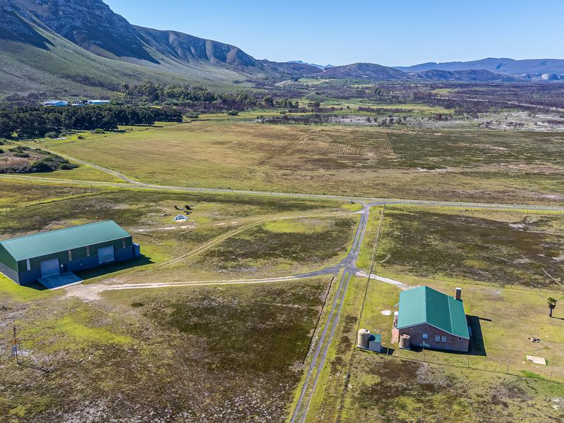Aerial view of a farm property featuring a large metal shed, a brick farmhouse with green roofing, and expansive surrounding agricultural land.
