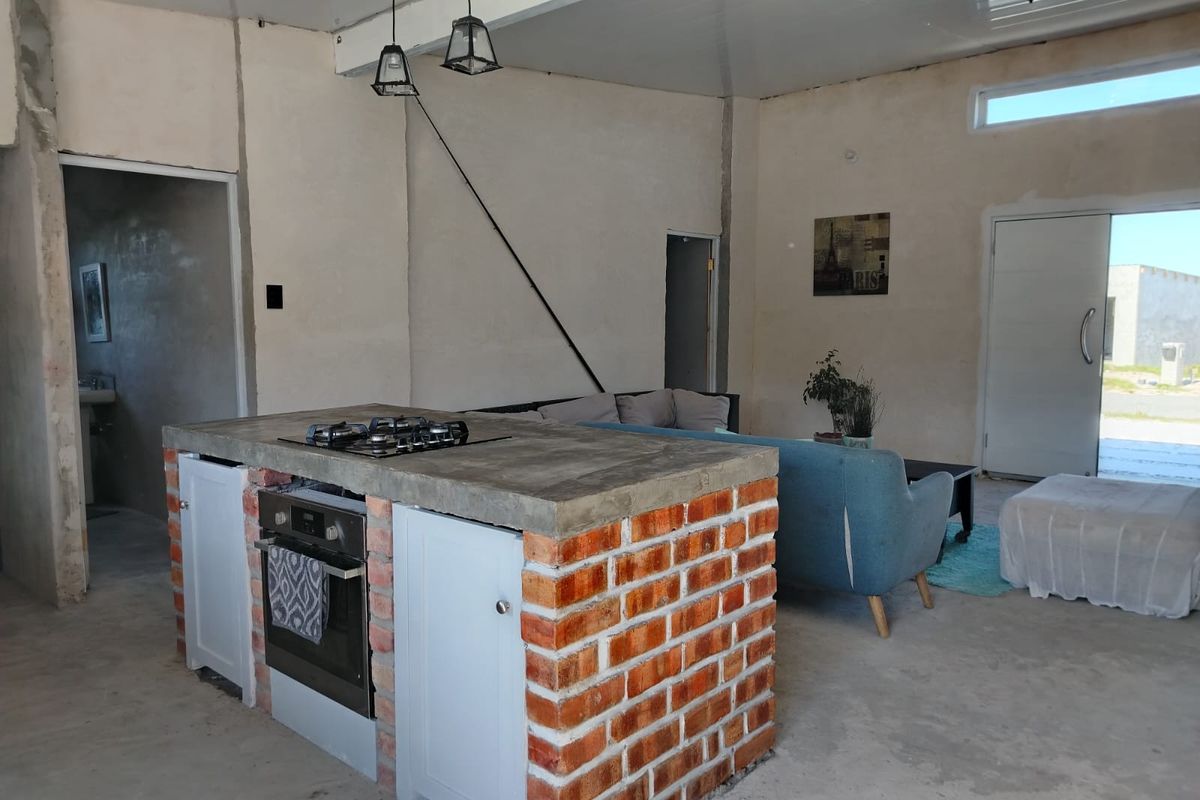 Open plan living area featuring an industrial-style kitchen island with exposed brick, concrete countertop, and built-in appliances, set within an unfinished interior space.