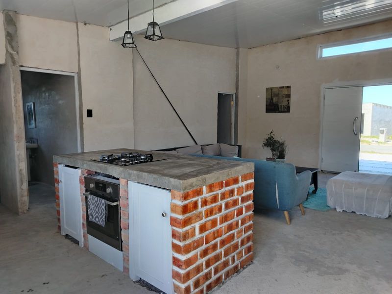 Open plan living area featuring an industrial-style kitchen island with exposed brick, concrete countertop, and built-in appliances, set within an unfinished interior space.