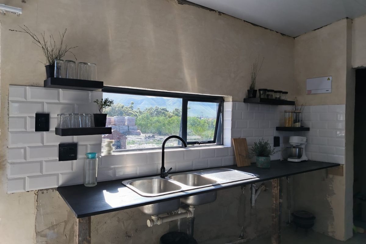 A kitchen area featuring a double bowl sink, subway tile backsplash, and floating shelves, currently requiring finishing touches to the walls and cabinetry.