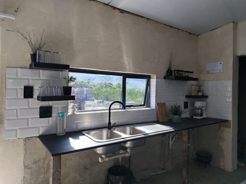 A kitchen area featuring a double bowl sink, subway tile backsplash, and floating shelves, currently requiring finishing touches to the walls and cabinetry.