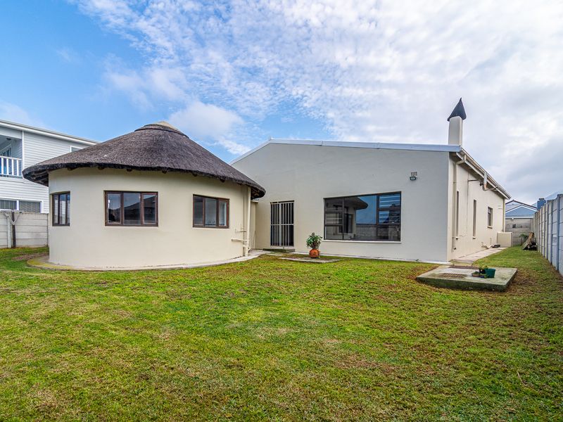 Rear view of the property featuring a unique circular structure with a thatched roof, a chimney, and a spacious grass garden area.