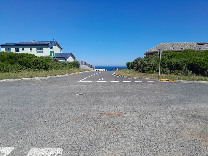 A clear street view showing the approach to the residential area with paved roads, kerbing, and distant ocean views.
