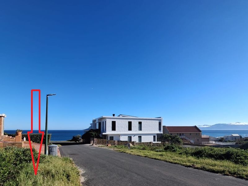 A street view of the residential area showing the road, coastal surroundings, and nearby properties under a clear blue sky.