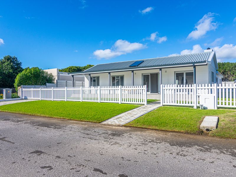 A well-maintained single-story house featuring a white facade, corrugated metal roof with solar panels, and a neat front garden with a picket fence.