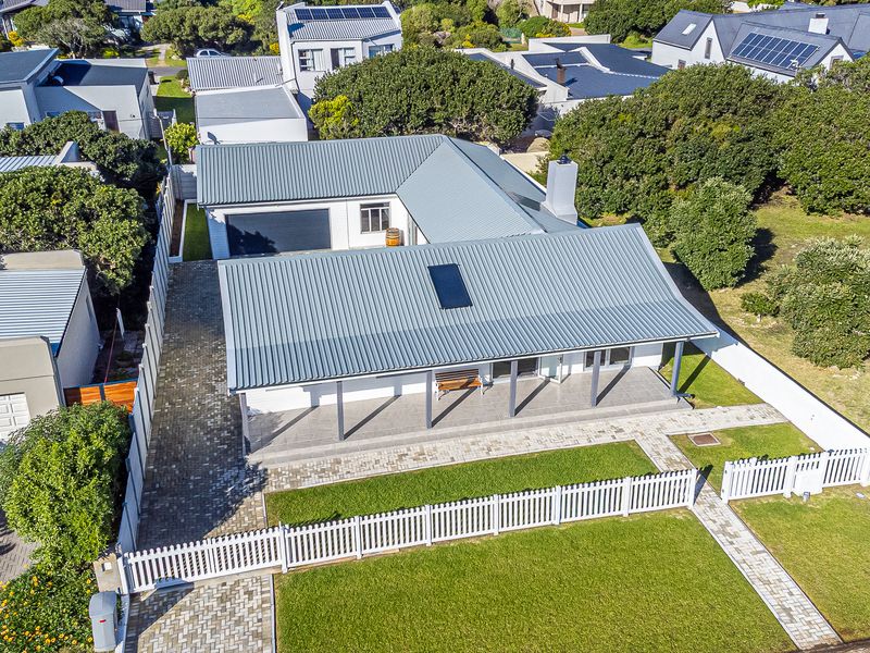 Aerial view of a well-maintained residential property featuring a metal roof, solar panel, covered patio, paved driveway, and a neat front garden with picket fencing.