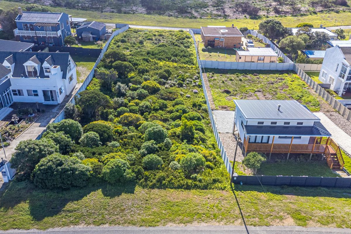 An aerial view of a vacant residential plot featuring natural vegetation and perimeter fencing, situated in a developed coastal neighborhood.