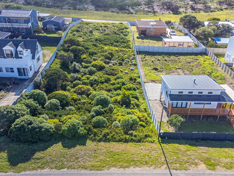 An aerial view of a vacant residential plot featuring natural vegetation and perimeter fencing, situated in a developed coastal neighborhood.