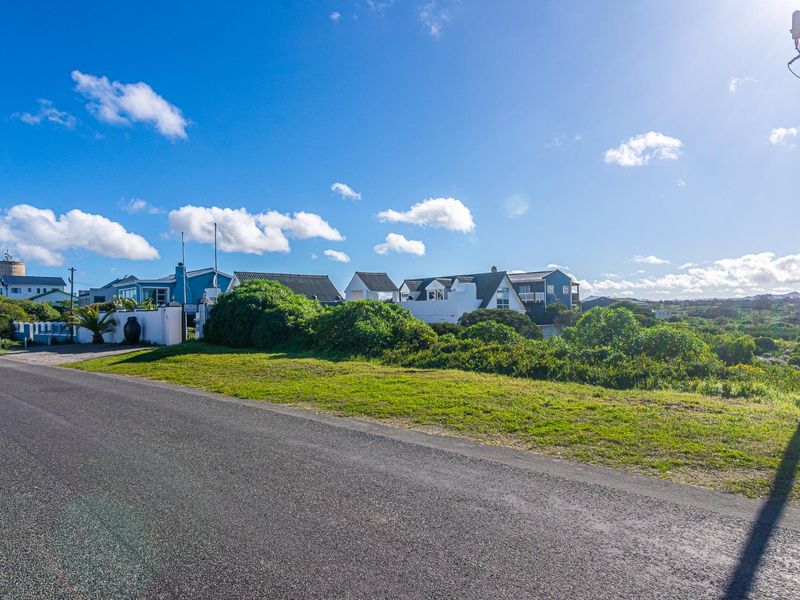 A street-level view of a residential area featuring an asphalt road, grass verge, and utility pole with surrounding coastal neighborhood homes in the background.
