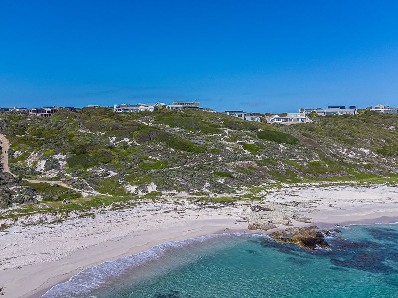 An aerial view of a pristine coastal landscape featuring a sandy beach, turquoise waters, and natural vegetation with residential properties situated on the hillside.