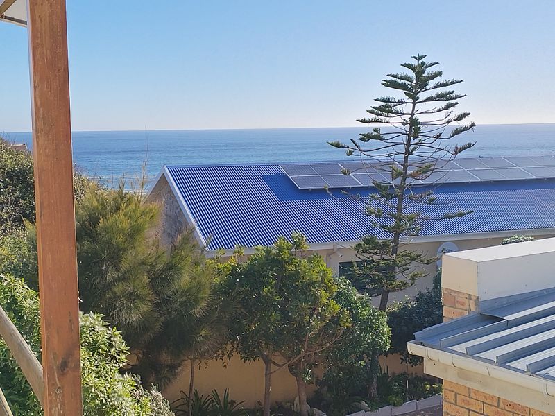 An elevated view overlooking a coastal property featuring a corrugated metal roof with integrated solar panels and a scenic ocean backdrop.
