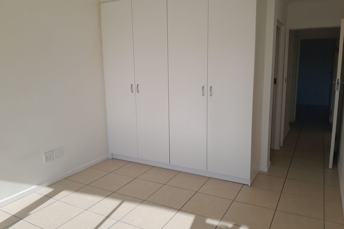 A bright, empty bedroom featuring neutral tiled flooring and ample built-in cupboard storage, leading into a hallway.