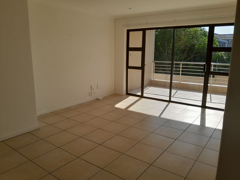 An empty, sunlit living room featuring neutral tiled flooring and large sliding glass doors that provide access to an outdoor balcony area.