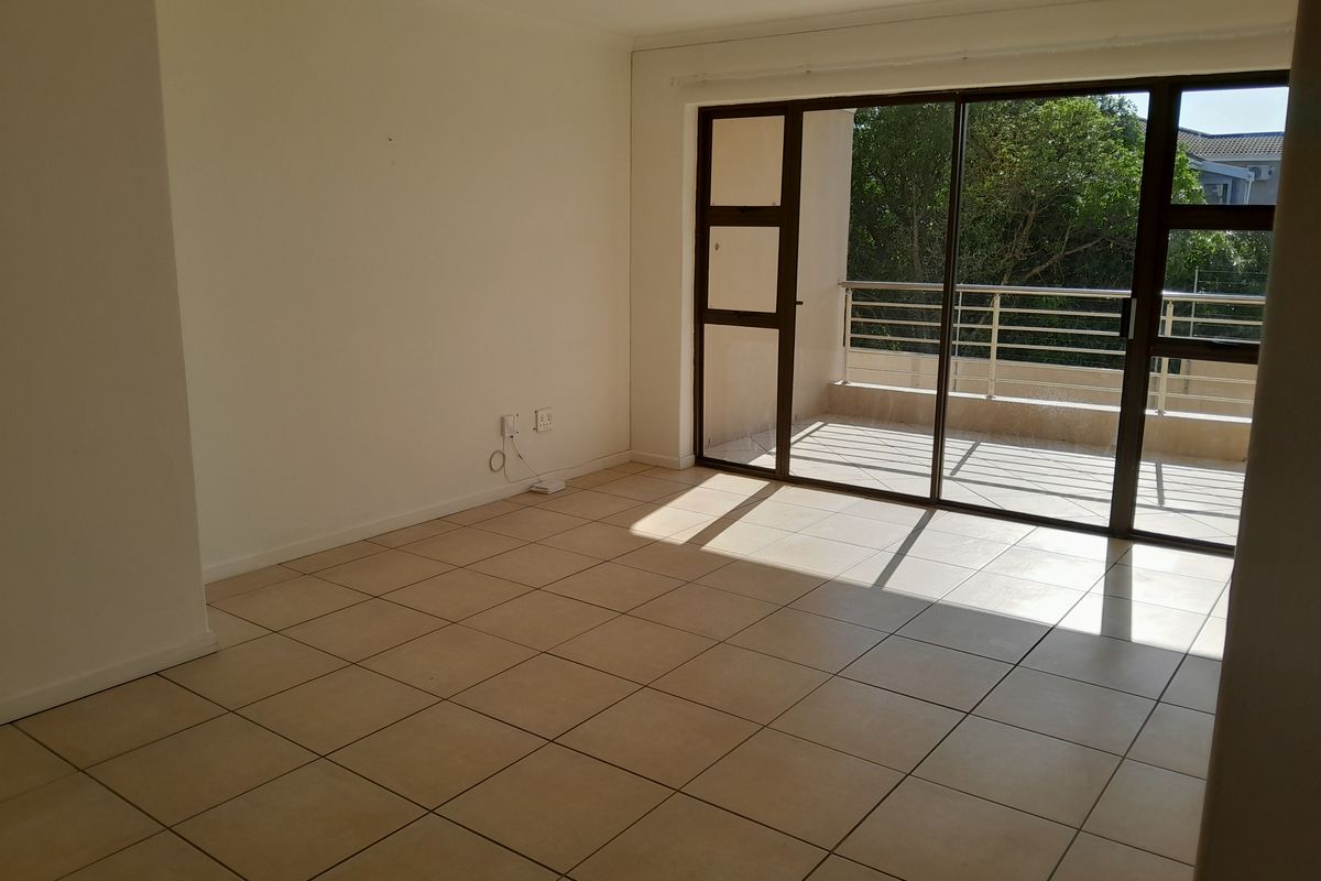 An empty, sunlit living room featuring neutral tiled flooring and large sliding glass doors that provide access to an outdoor balcony area.