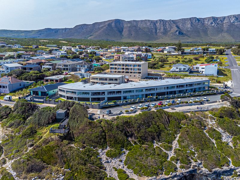 Aerial view of a modern apartment complex situated on a coastal cliff, featuring extensive parking and proximity to the surrounding residential neighborhood.