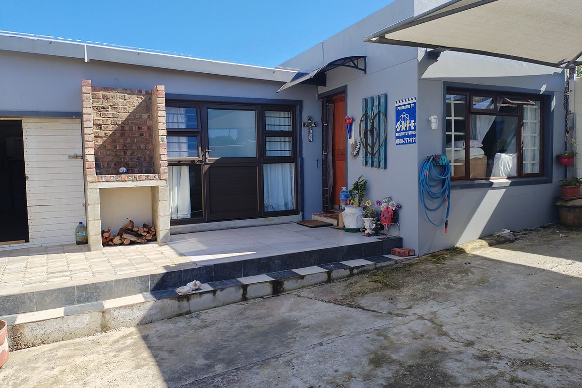 Rear exterior view featuring a tiled patio area with a built-in brick braai, aluminium window frames, and a concrete driveway.