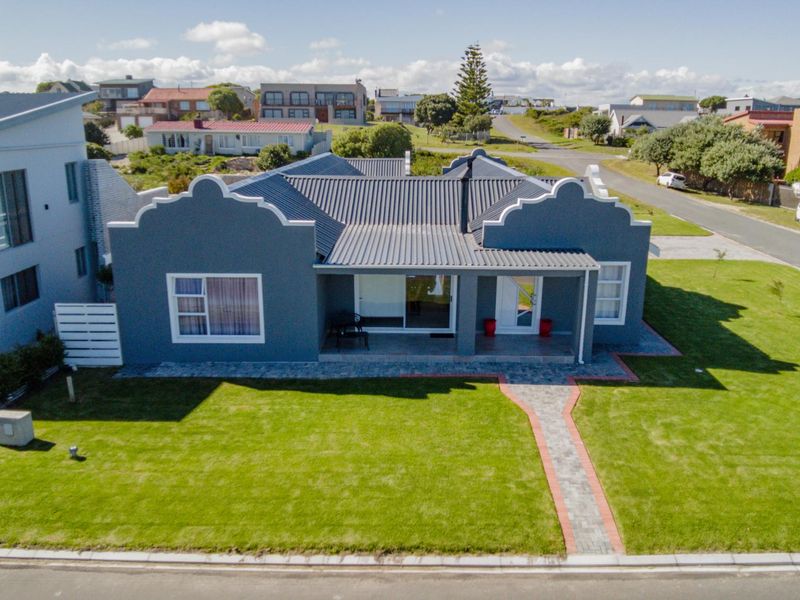 Aerial view of a well-maintained single-story house featuring a grey facade, corrugated metal roof, and a neat front lawn with a paved walkway.