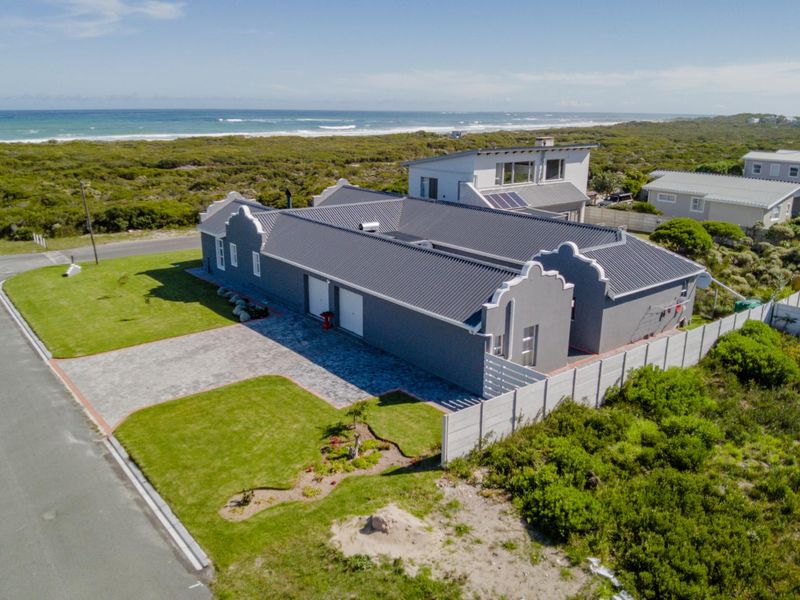 Aerial view of a coastal property featuring a modern grey house with a metal roof, paved driveway, and secure boundary wall near the ocean.